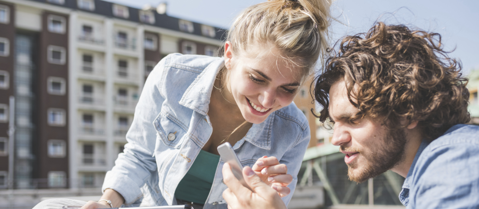 Mosolygó fiatal barna hullámos hajú férfi és szőke nő nézik a mobiltelefonjukat a szabadban.