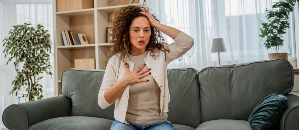 Stresszes középkorú nő ül a kanapén, és küzd a mellkasi fájdalommal és a légzési nehézségekkel otthon.