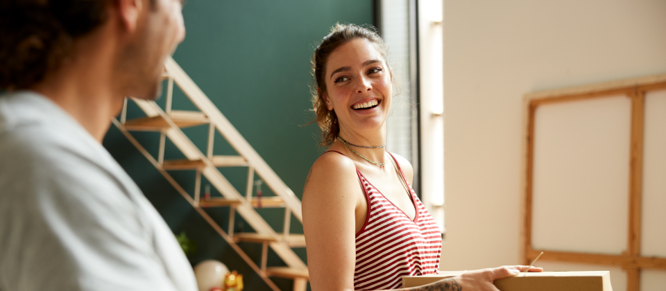 Egy lány mosolyogva néz a párjára és egy dobozt tart a kezében az új otthonukban.